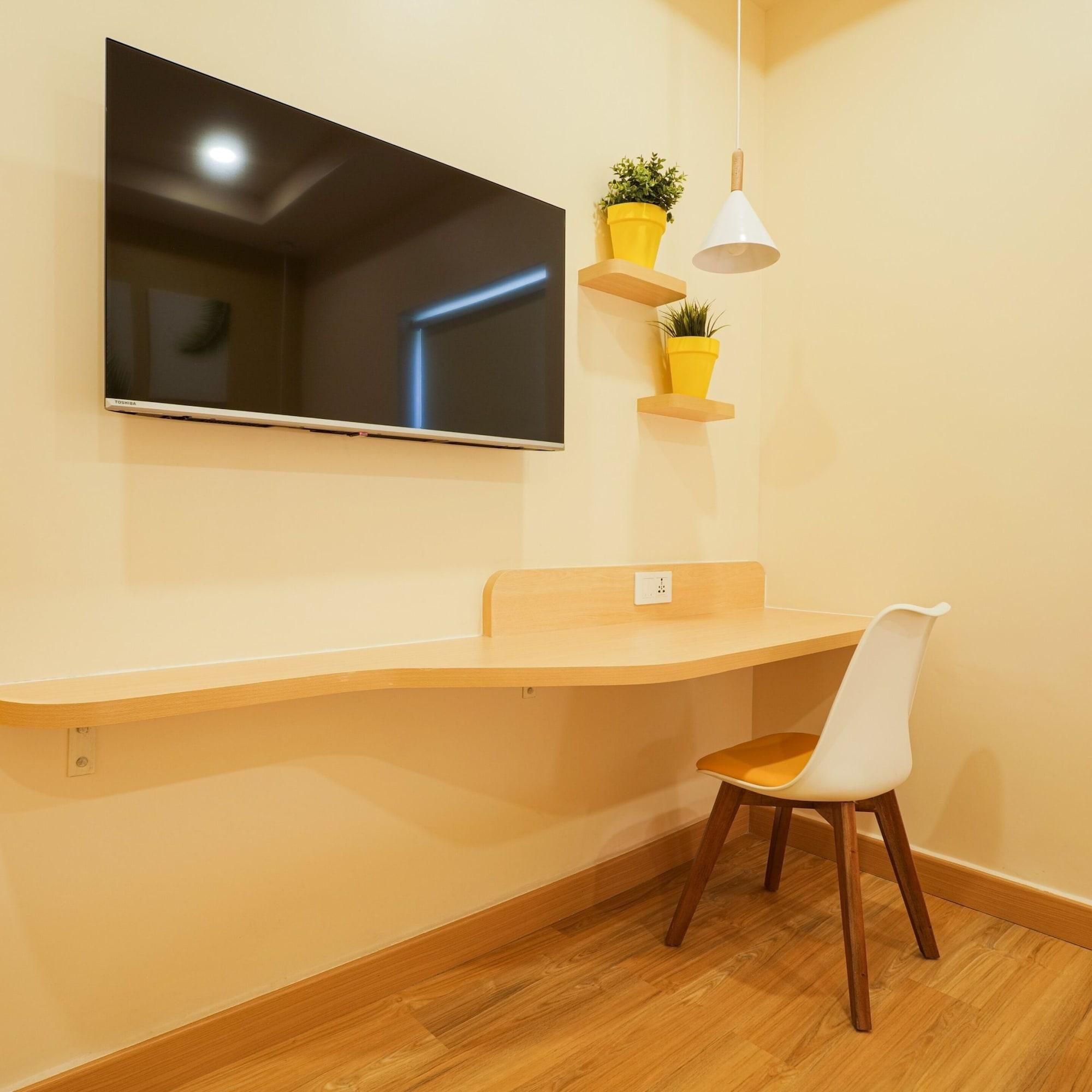 A modern room with a flat-screen TV mounted on the wall, a wooden desk with a chair, and two potted plants on floating shelves.