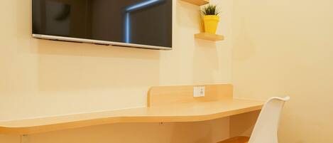 A modern room with a flat-screen TV mounted on the wall, a wooden desk with a chair, and two potted plants on floating shelves.