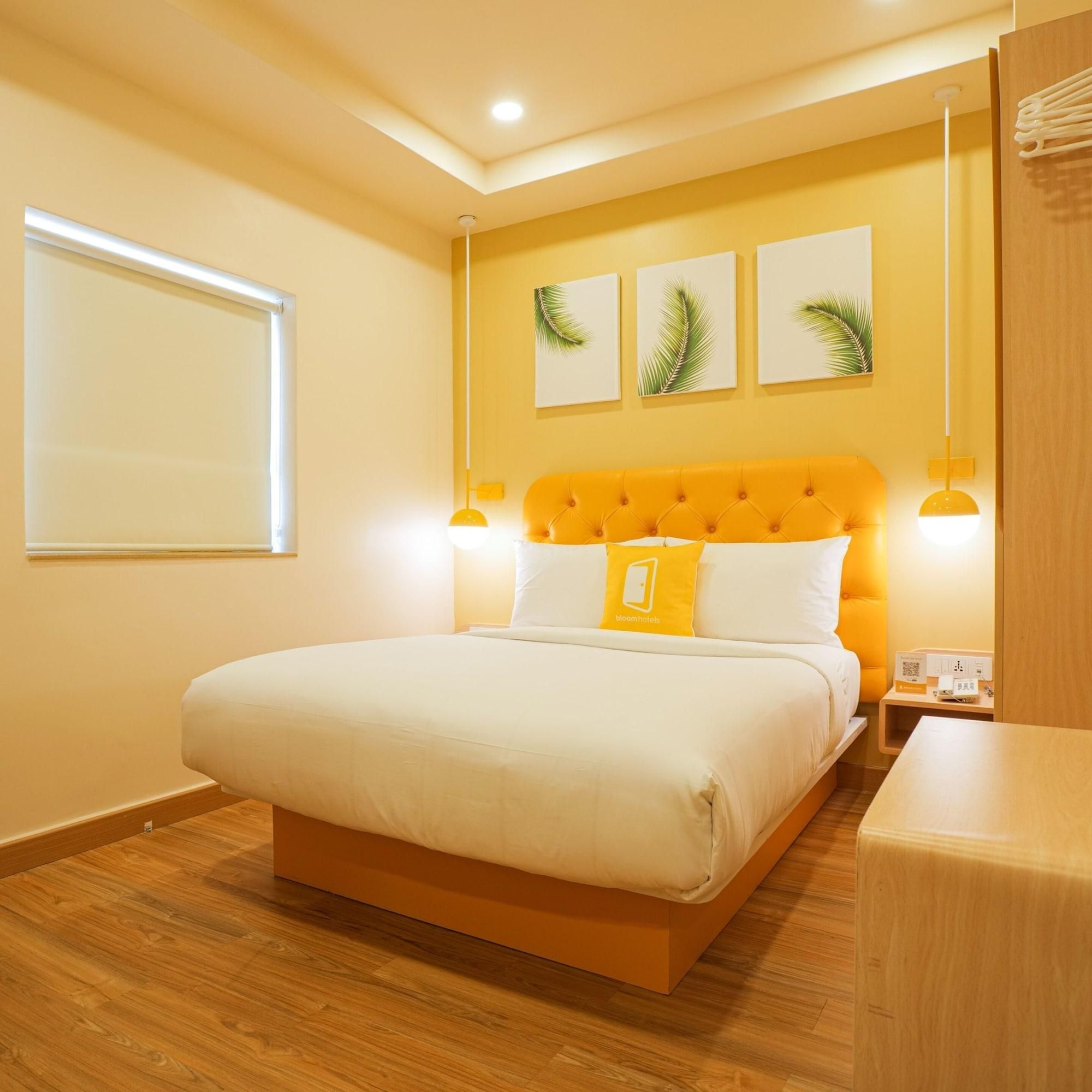 A hotel room with a large bed, a wooden floor, a window with blinds, and a wall with three framed palm leaf pictures.