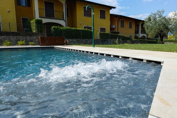 A swimming pool with clear blue water, surrounded by a concrete deck and a yellow building with balconies in the background. - Gardaliva 6 by the lake in Manerba del Garda (Manerba del Garda)