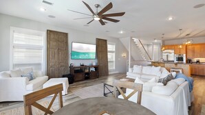 A spacious living room with a large TV, a dining area, and a kitchen in the background.