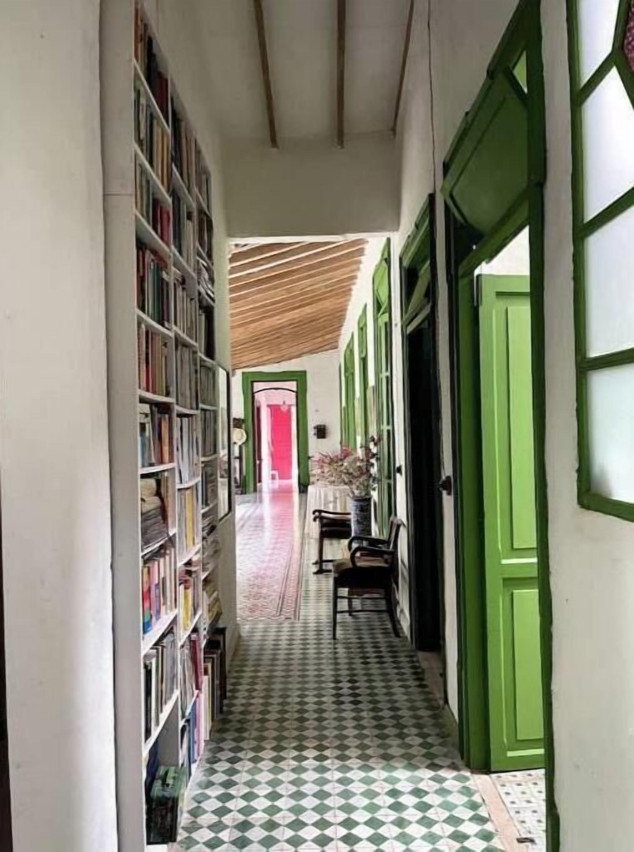Um corredor estreito com chão quadriculado, portas verdes e uma estante de livros em um dos lados.