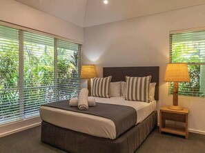 Ein Schlafzimmer mit einem großen Bett, zwei Nachttischlampen und Blick auf Grün durch die Fenster.