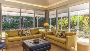 Ein geräumiges Wohnzimmer mit zwei gelben Sofas, einem dunklen Hocker und einem großen Fenster mit Blick auf üppiges Grün.