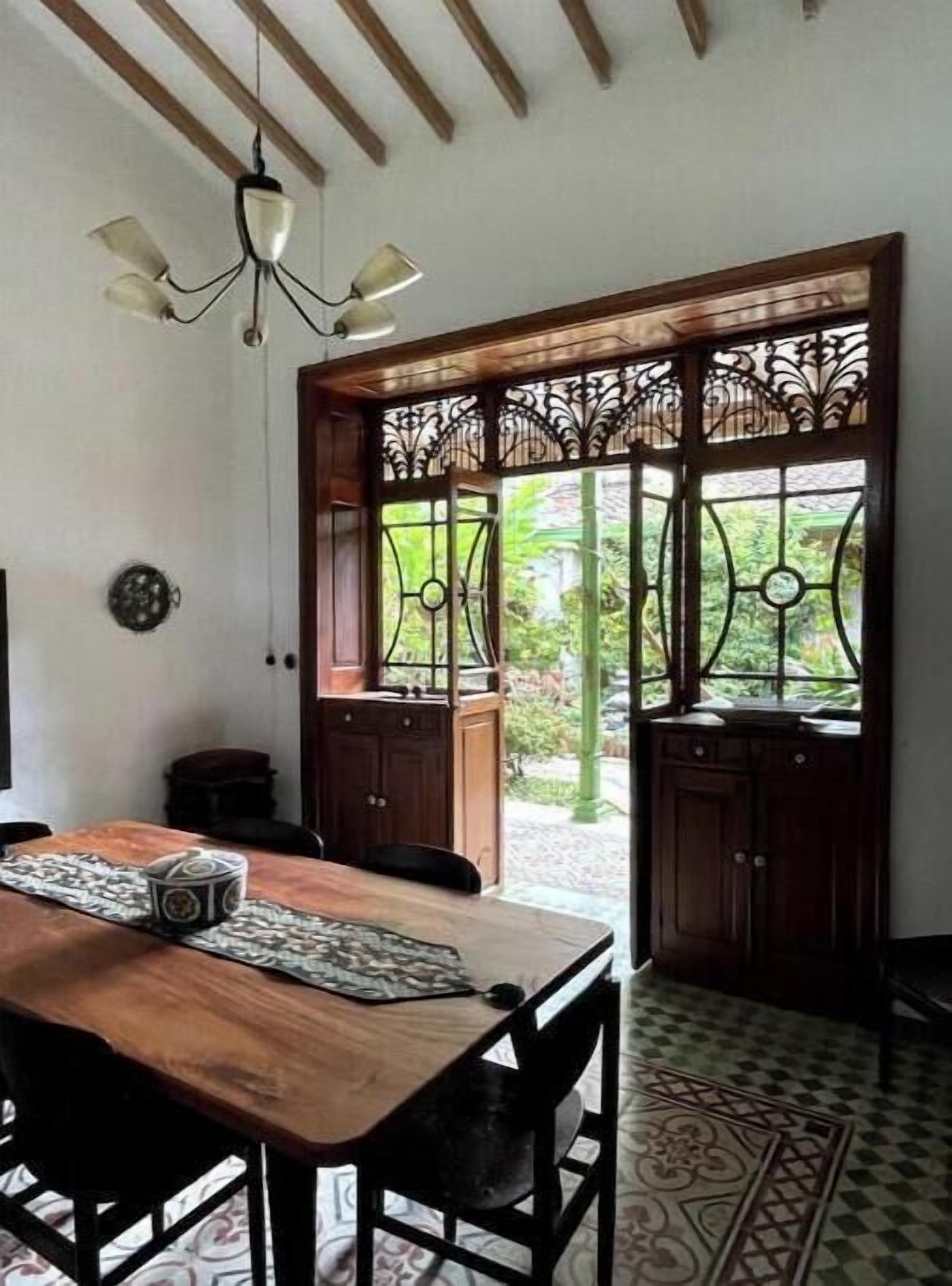 Un coin repas avec une table et des chaises en bois, un ventilateur de plafond et une vue sur un jardin, visible à travers une porte décorative.