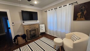 A living room with a fireplace, a television mounted above it, a white armchair, a striped rug, and a ceiling fan. - Easy Living In Delightful Duplex (Lubbock)