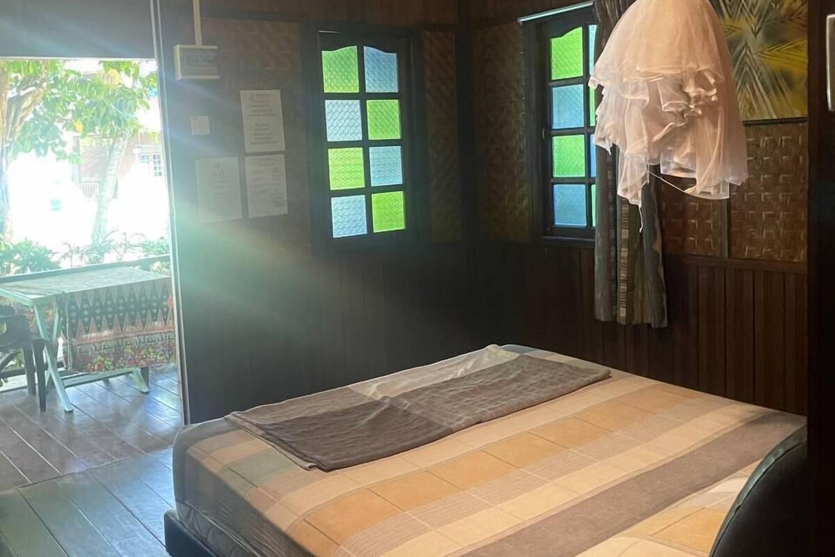 A room with a bed, a ceiling fan, a mosquito net, and a window with a view of greenery.