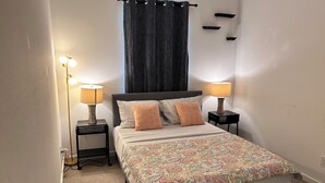 Un dormitorio ordenado con cama, dos mesitas de noche, dos lámparas, una cortina oscura y una colcha estampada.