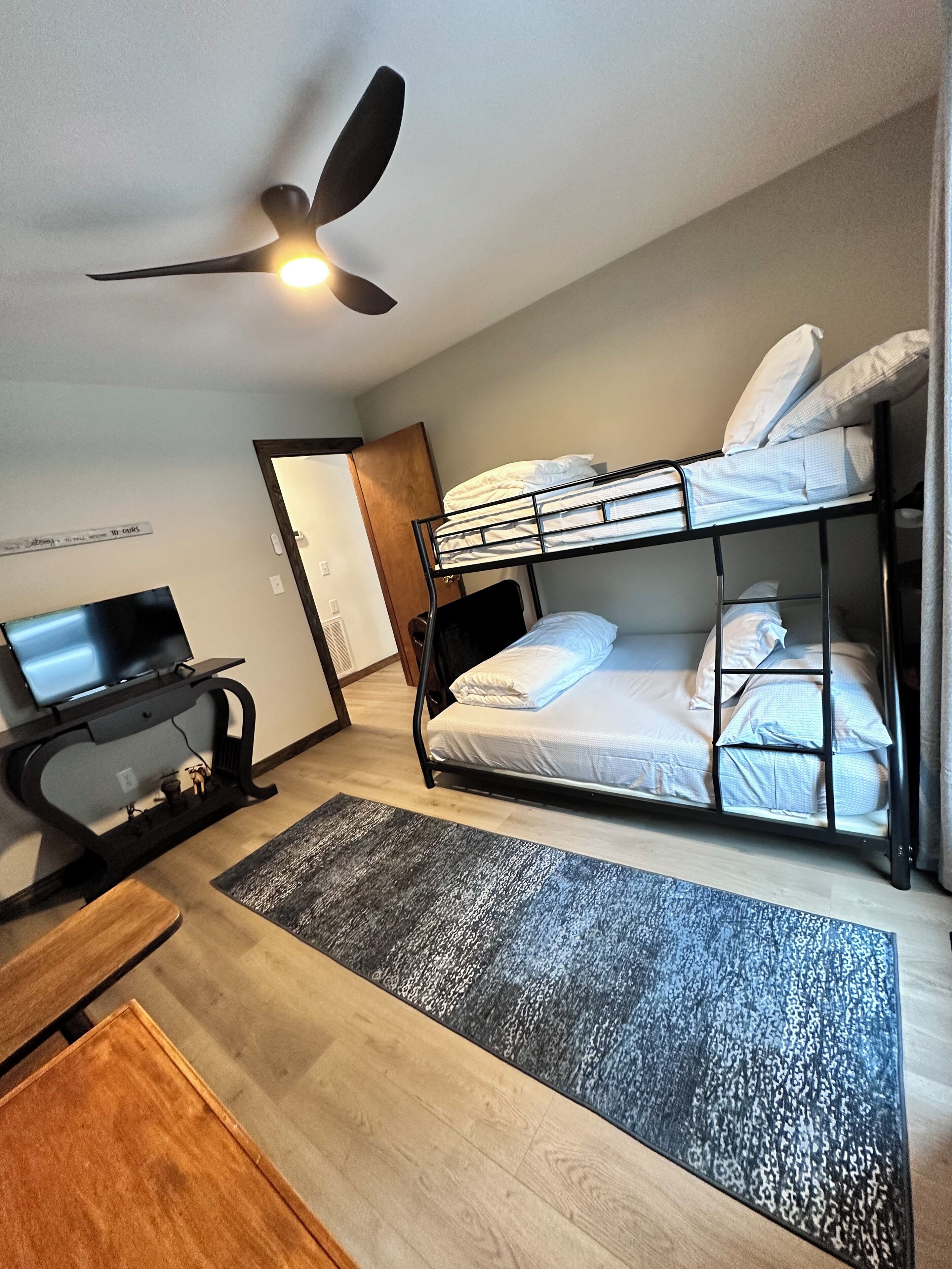 Une chambre d’hôtel compacte avec des lits superposés, un bureau avec un ordinateur, un ventilateur de plafond et un petit tapis.