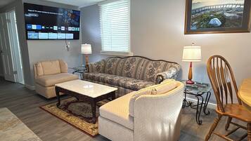 Un salon avec un canapé beige, un canapé à motifs, une table basse, une petite table avec une lampe et un téléviseur fixé au mur.
