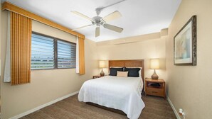 Une chambre d’hôtel comprenant un lit, deux tables de chevet avec des lampes, un ventilateur de plafond, une fenêtre avec des stores et un tableau encadré au mur.