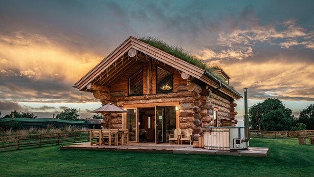Una cabaña de troncos con techo de hierba, ventanas amplias y un patio con mobiliario exterior.