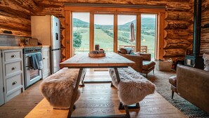 Interior rústico de una cabaña con una mesa de madera, bancos, una zona de cocina y un amplio ventanal con vistas al campo.