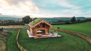 Una cabaña de troncos con techo verde, rodeada por una valla de madera, y con un patio que incluye mobiliario exterior y un jacuzzi.