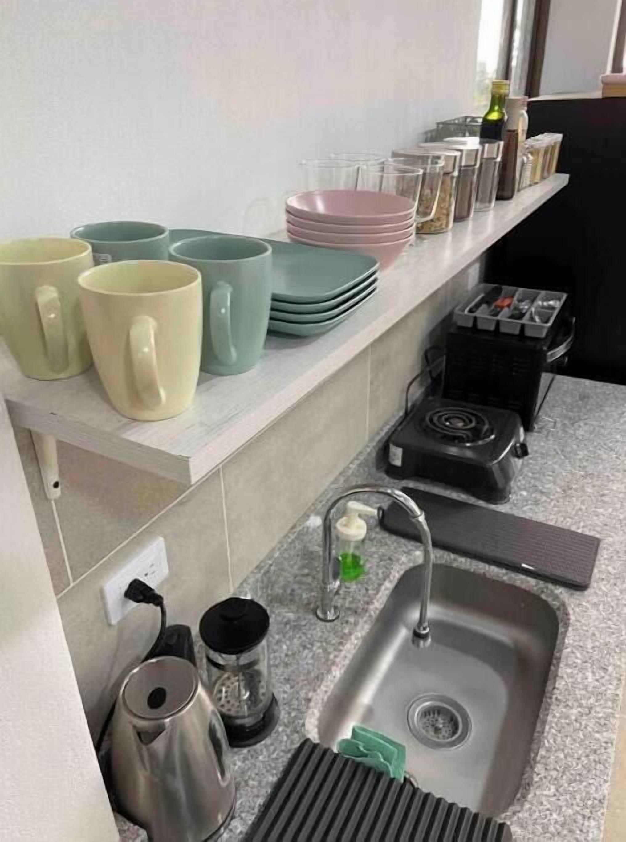 Uma cozinha com pia, chaleira e cafeteira. Há pilhas de pratos e xícaras sobre o balcão.