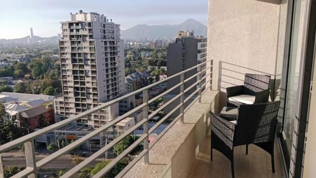 A balcony with a table and chairs, overlooking a cityscape with buildings and mountains in the distance.