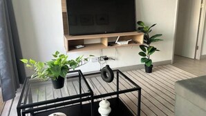 A modern living room with a flat-screen TV mounted on a wooden shelf, a glass coffee table with decorative items, and potted plants.