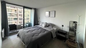 A modern bedroom with a large bed, a small bedside table, a mirror, and a view of a building through a sliding glass door.