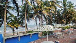 Eine Terrasse direkt am Strand mit Tischen und Stühlen, Palmen und Blick auf das Meer.