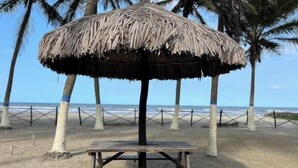 Ein Strandsonnenschirm mit einer hölzernen Sitzbank darunter, umgeben von Palmen und einer umzäunten Fläche.