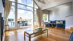 A spacious living room with a wooden floor, a blue sofa, and a glass coffee table. Large windows offer a view of the outdoors. - The Landings 18, Waters Reach (P) - Lakeside Luxury, Hot Tub, Tennis, Sleeps 10 (South Cerney)