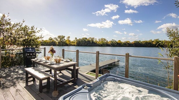 A wooden deck with a hot tub, picnic table, and grill overlooking a lake. - The Landings 18, Waters Reach (P) - Lakeside Luxury, Hot Tub, Tennis, Sleeps 10 (South Cerney)