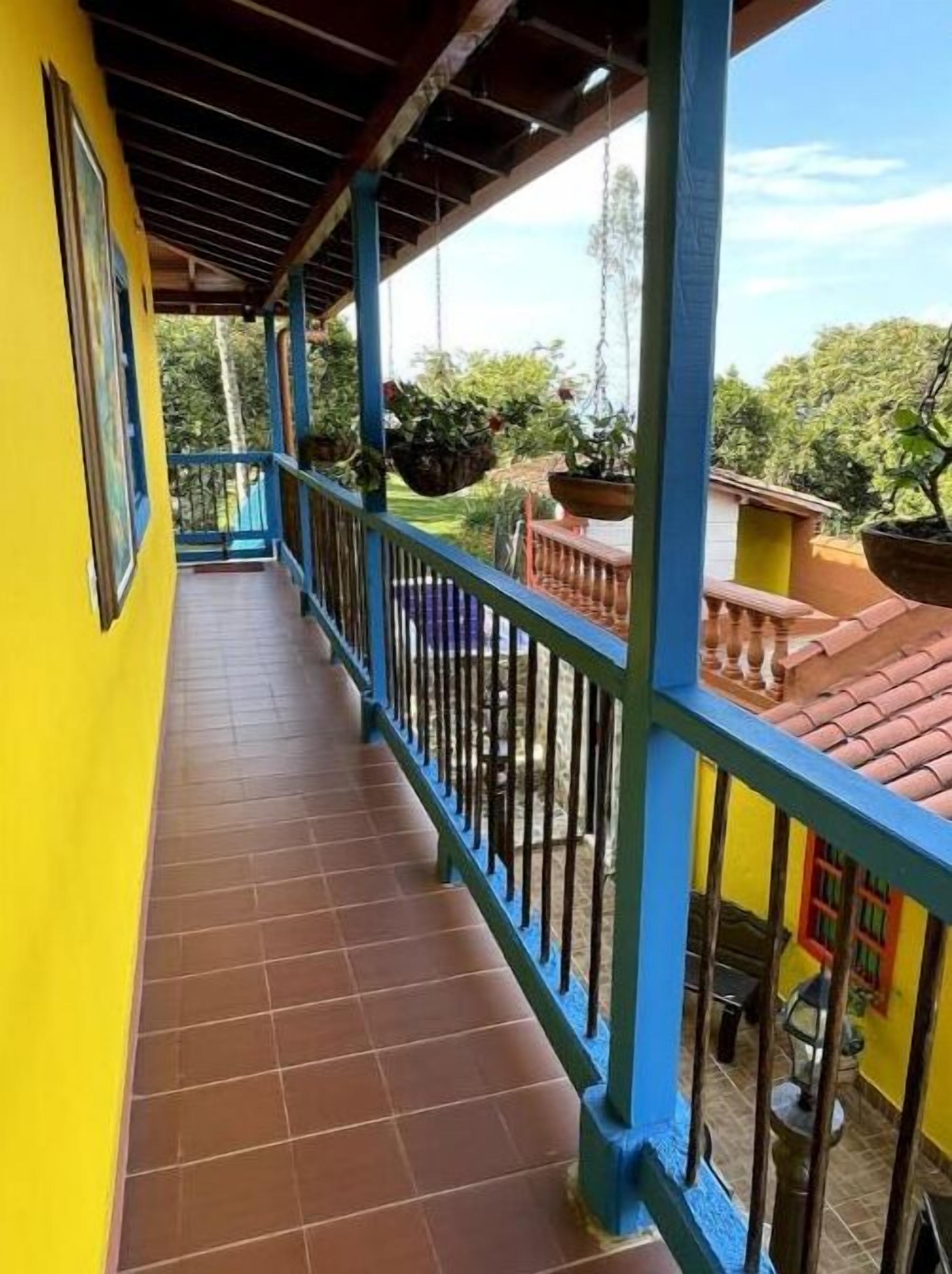 A covered balcony with blue railings, a tiled floor, and a view of greenery and buildings in the background.