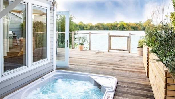 Een houten terras met een jacuzzi, een glazen deur die leidt naar een overdekt gedeelte en uitzicht op een meer.