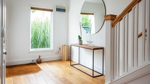 Een kamer met houten vloer, een witte deur, een spiegel, een console met een plant, en een raam met uitzicht op groen.