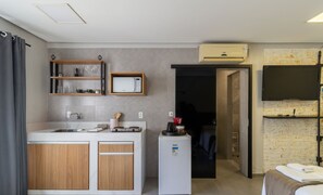 A compact kitchen with a built-in fridge, a microwave, and a coffee machine. There is a wooden cabinet and a shelf with glassware. - Estúdio Modernos em Condomínio com Piscina em Floripa - ASK (Florianópolis)