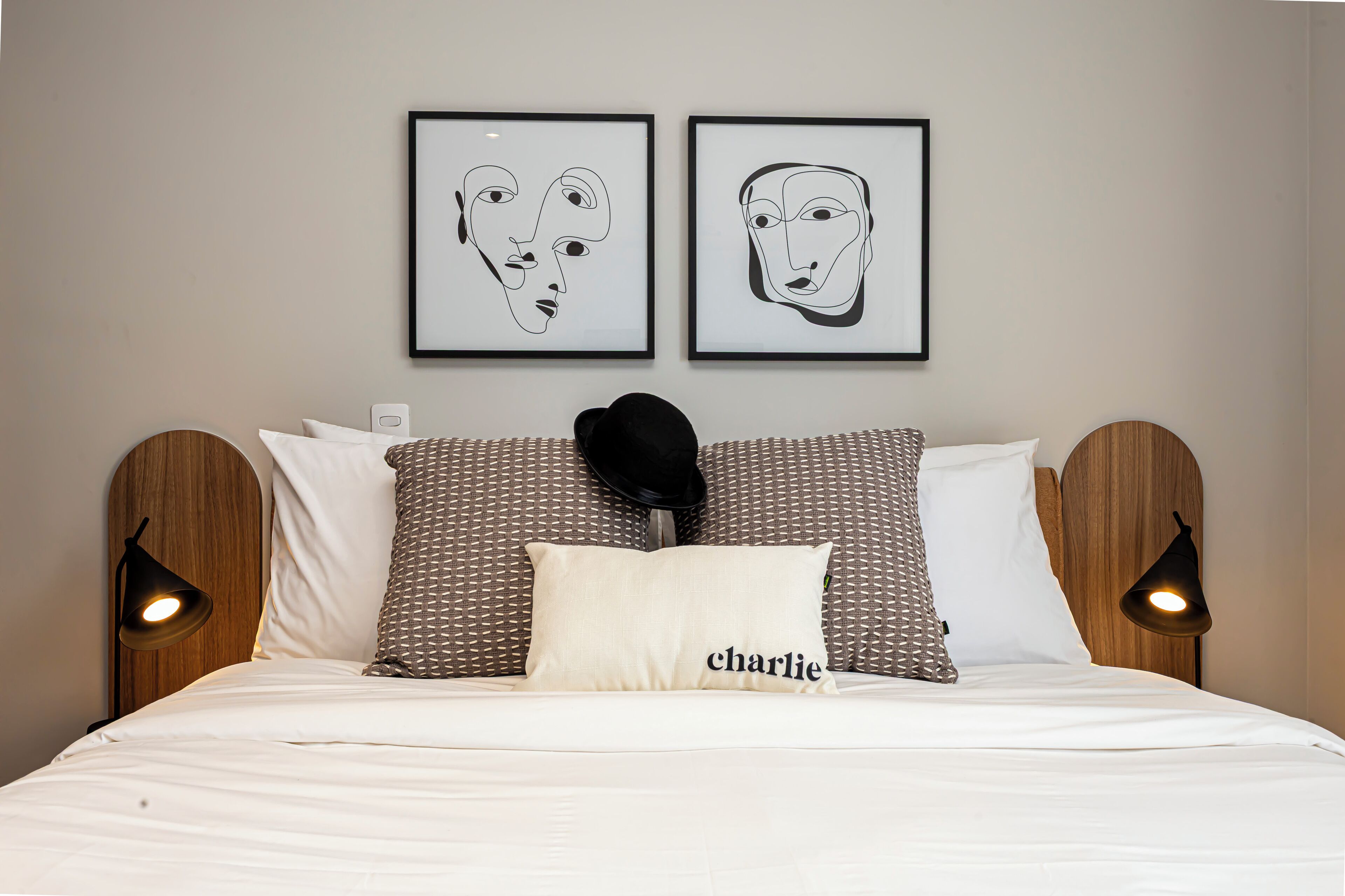 A neatly made bed with a pillow labeled 'charlie,' two framed artworks on the wall, and two bedside lamps.