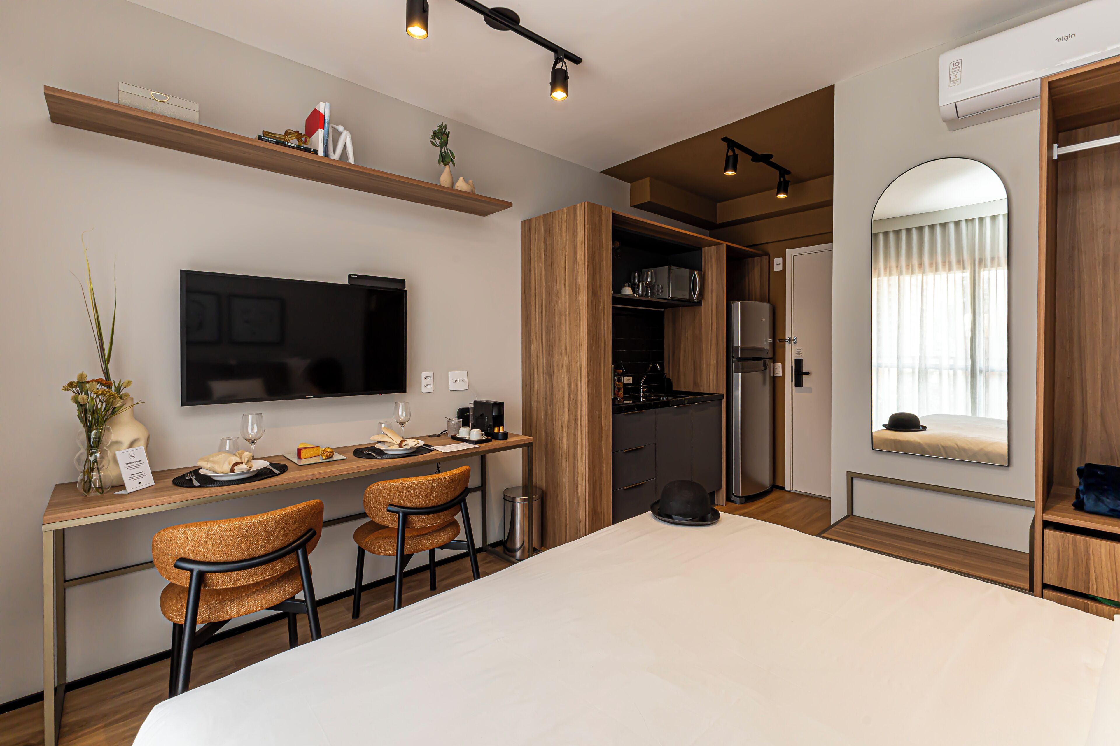 Quarto de hotel moderno com uma cama, uma mesa com duas cadeiras, uma TV, micro-ondas e uma pequena copa.