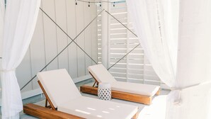 Deux fauteuils de jardin en bois, dotés de coussins blancs, sont placés sous des rideaux blancs, avec une lanterne blanche entre les deux.