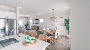 Une cuisine moderne dotée d’un four encastré, un coin repas avec une table en bois et des chaises en osier, et un salon avec un canapé et un téléviseur.