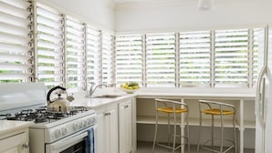 Eine Küche mit weißen Schränken, einem Herd und einem Spülbecken. Zwei Barhocker und ein Fenster mit Jalousien sind vorhanden.