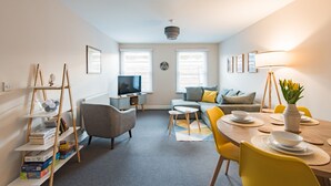 A modern living room with a sofa, armchairs, a dining table set for four, a TV, and a bookshelf.