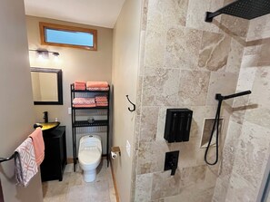 Une salle de bain avec des toilettes, un lavabo et une douche. On y trouve une étagère avec des serviettes et un miroir au-dessus du lavabo.