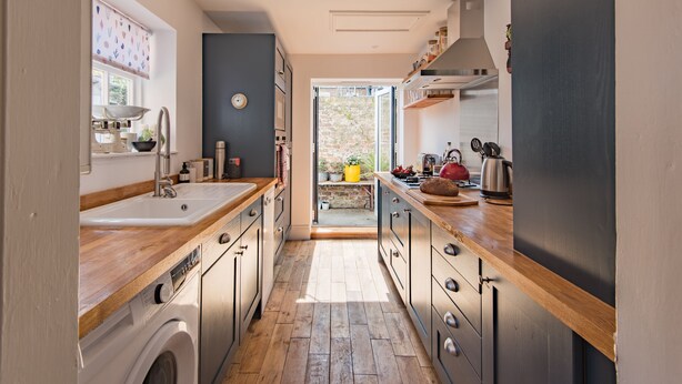 A narrow kitchen with wooden countertops, a washing machine, and a view to an outdoor area with a brick wall and plants. - Tell-Tale Cottage, Deal (Deal)