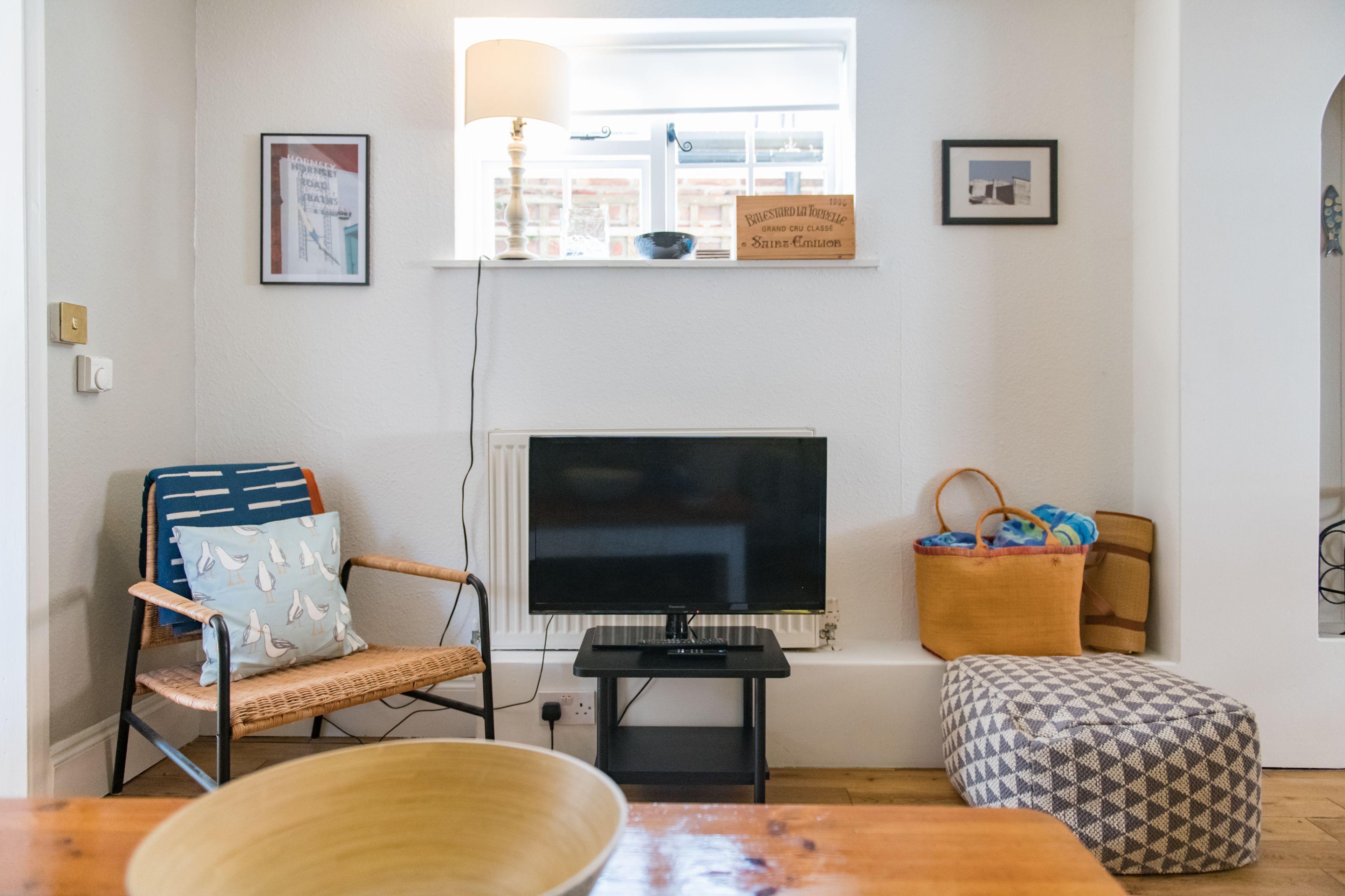 A living room with a television, a wicker chair, a wooden table, and a patterned ottoman.