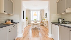 A kitchen with wooden flooring, white cabinets, a dining table with chairs, and a hanging pendant light. - Michaelmas Cottage, Broadstairs (Broadstairs)