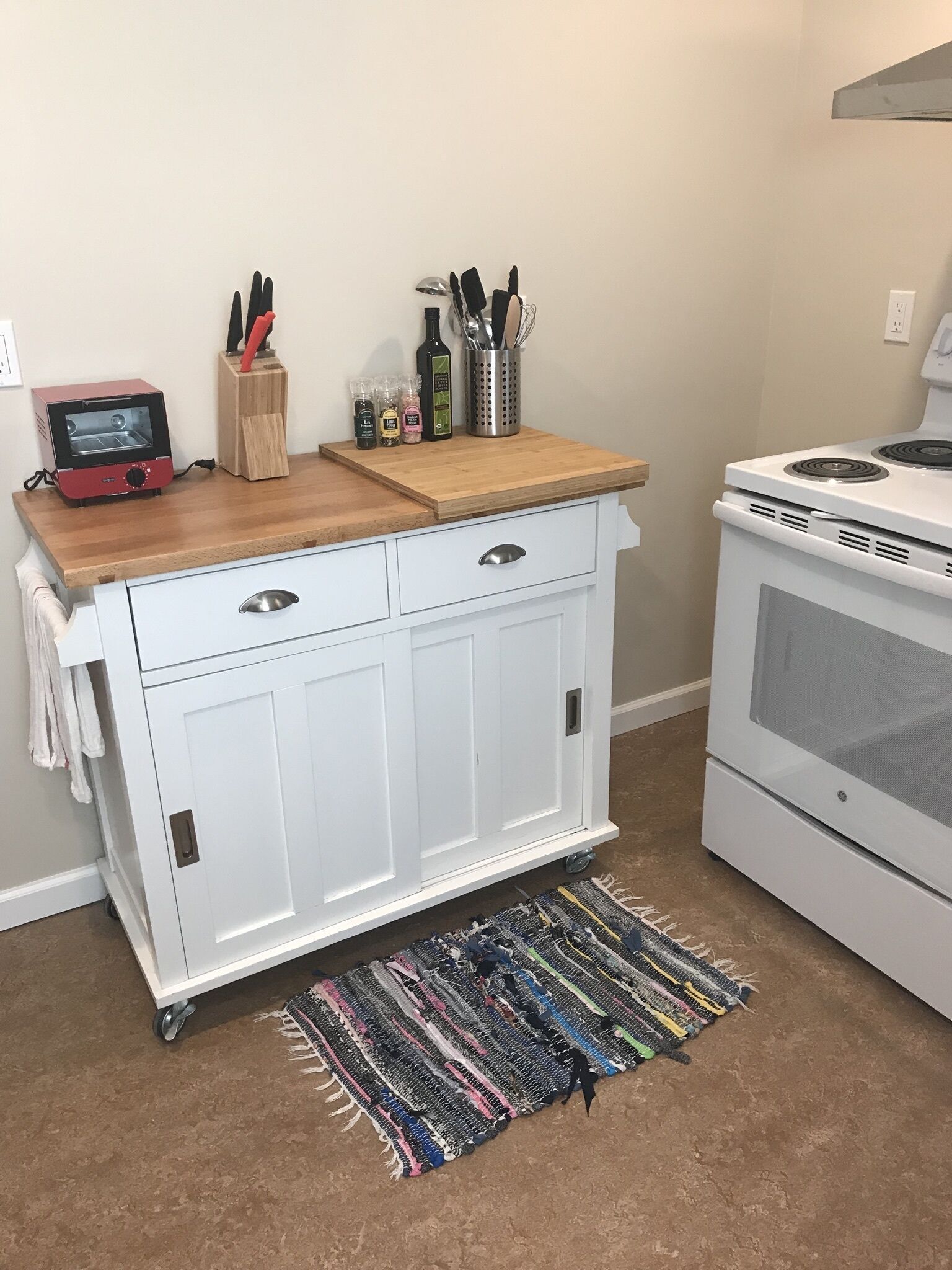 A kitchen with a white cabinet, a red toaster, a stove, and a colorful rug on the floor.