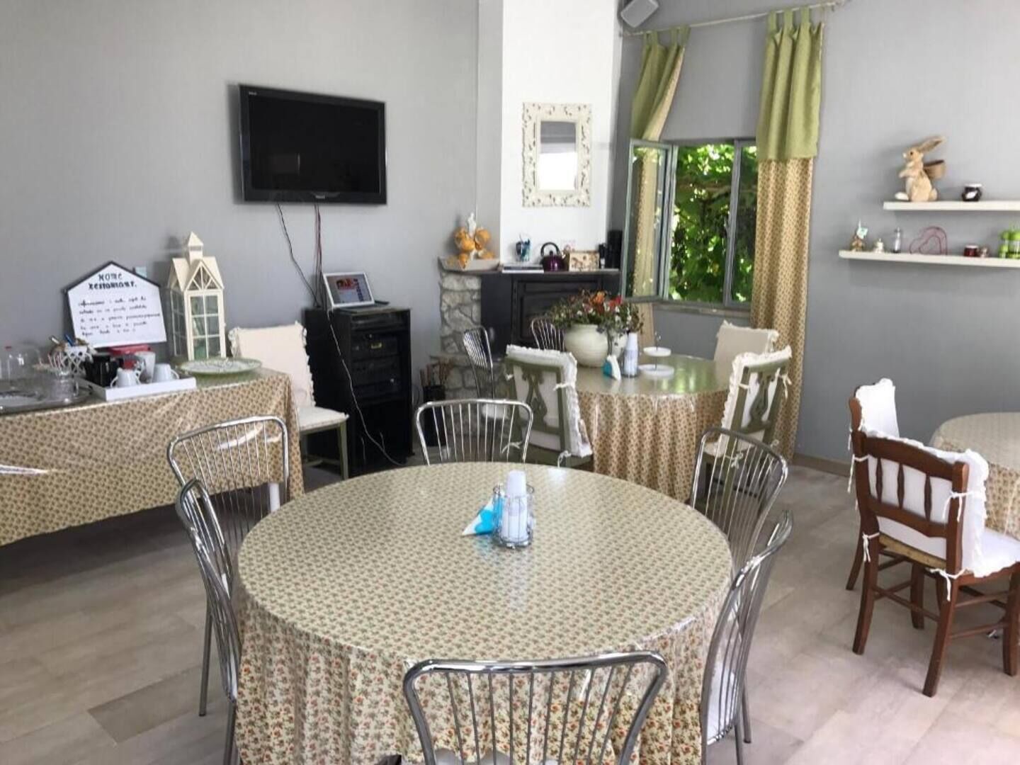 Ein Essbereich mit runden Tischen, die mit floralen Tischdecken bedeckt sind, Metallstühlen und einem an der Wand befestigten Fernseher.