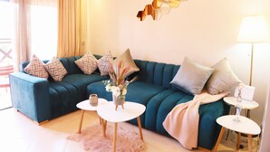 Ein petrolfarbenes Sofa mit gemusterten Kissen, ein kleiner weißer Tisch mit Blumen in einer Vase und eine Lampe auf einem Beistelltisch.
