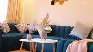 Ein blaugrünes Sofa mit gemusterten Kissen, ein kleiner weißer Tisch mit einer Vase gefüllt mit Trockenpflanzen und eine geometrische Wandleuchte.