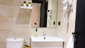 Ein Badezimmer mit einem weißen Waschbecken, einem Spiegel, einem Föhn und einem Regal mit einer Pflanze und einer Flasche.