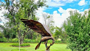 Una escultura de pájaro sobre un tocón de árbol, rodeada por un borde de piedras y plantas verdes en un jardín.