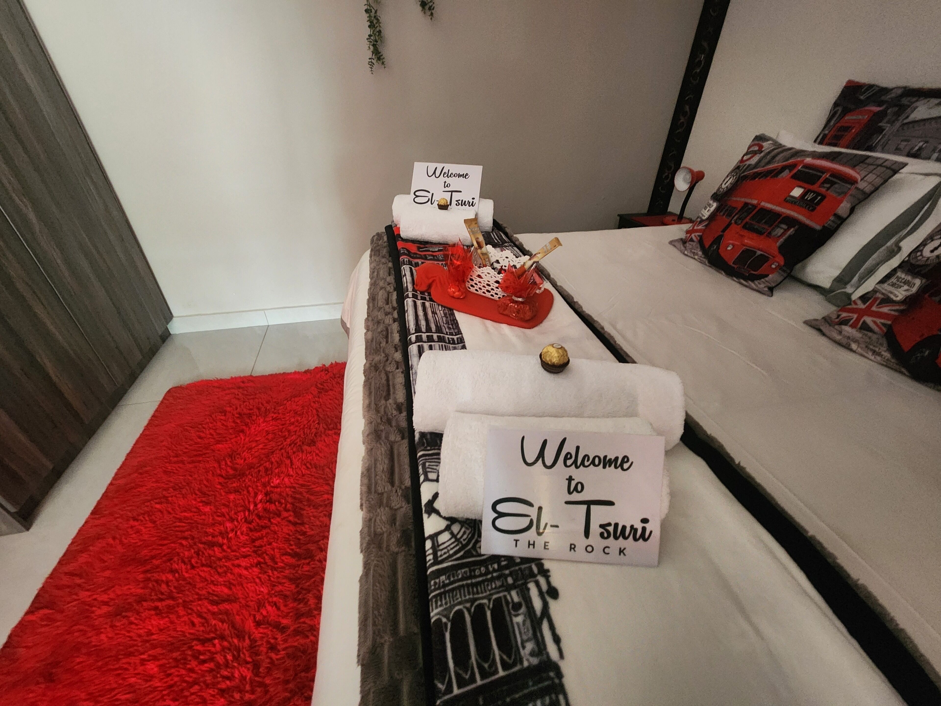 Un letto con una coperta rossa, un cartello con la scritta 'Welcome to El-Tsuri THE ROCK' e un cuscino a forma di autobus rosso.