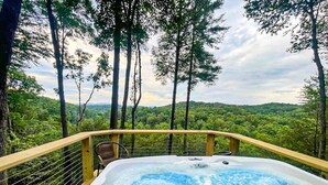 Un jacuzzi con cubierta de madera y baranda, rodeado de árboles y un paisaje boscoso.