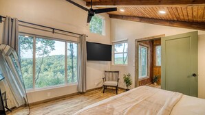 Un dormitorio con una cama grande, una silla, un televisor y vistas al bosque a través de amplios ventanales.