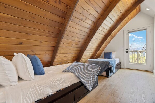 A cozy attic bedroom with wooden slats on the ceiling, two beds with pillows, and a door leading to a balcony.
